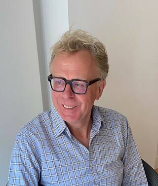 Colour photograph of man in blue shirt wearing black-framed glasses